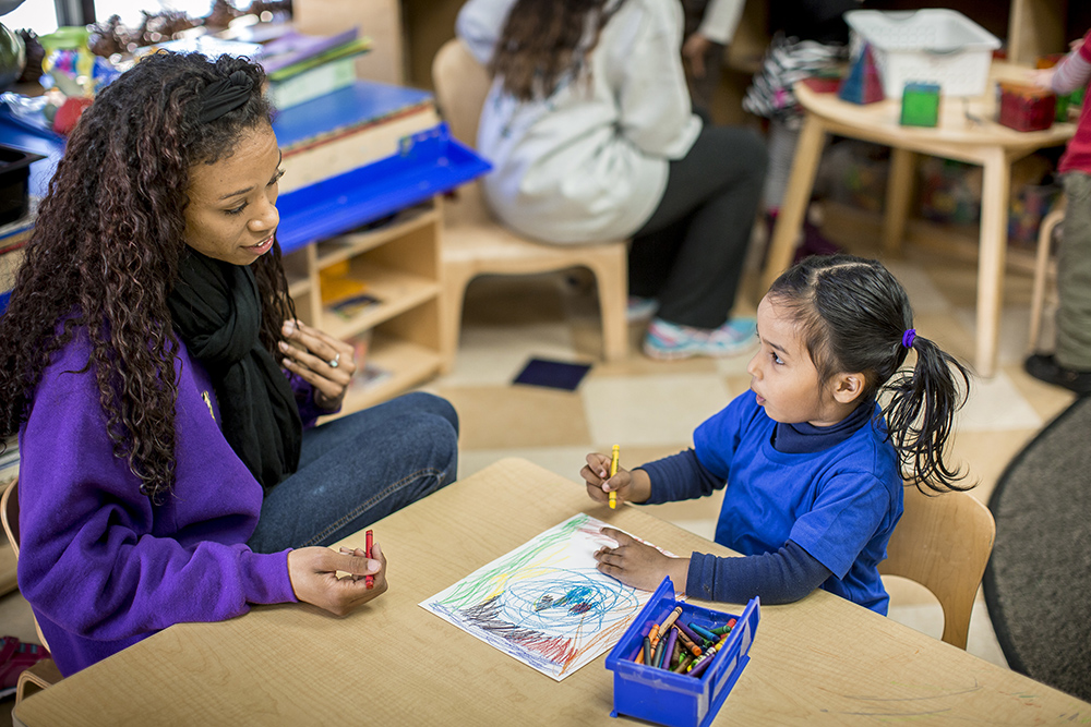 Female teacher helping a young girl color a picture.