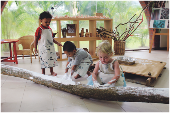 Three young children dressing in aprons using their hands to pait a tree branch white.