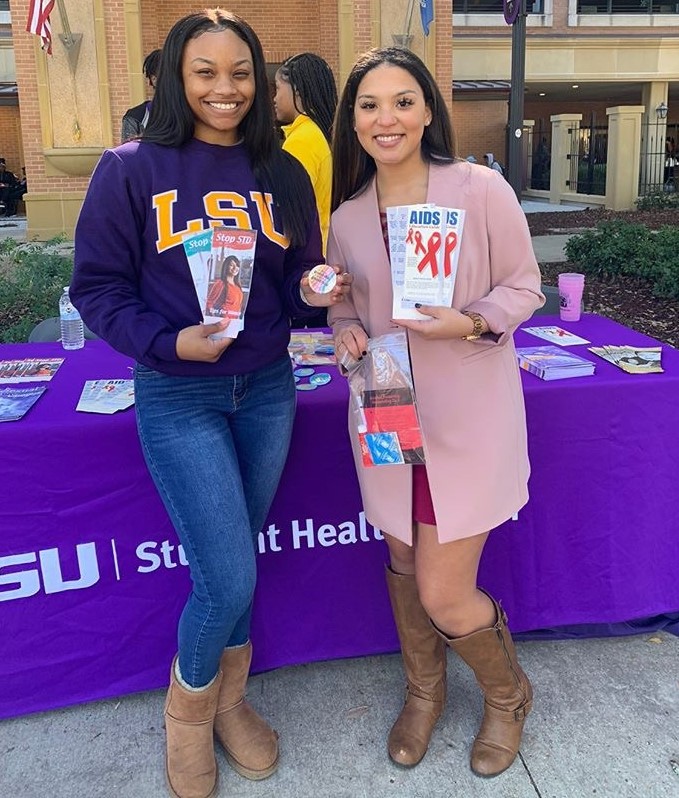 Black female student and Alaysia smile together in front of the LSU Student Health purple table cloth holding AIDS pamphlets.