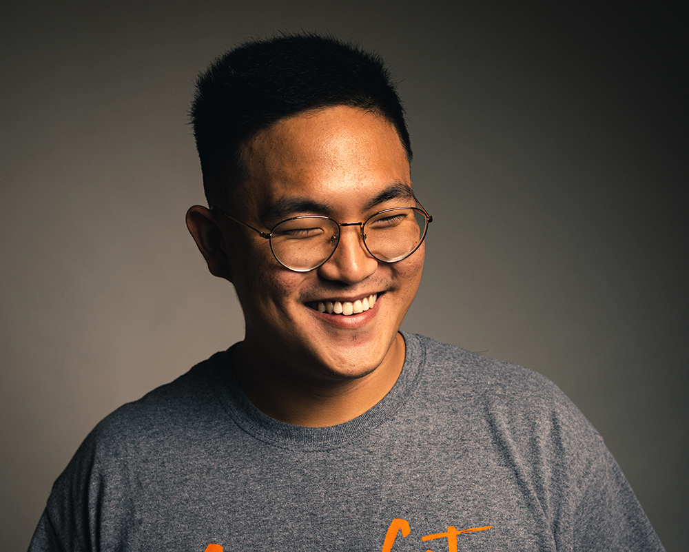 Joseph Park, an interdisciplinary major with minors is photography and sociology, smile while looking off screen. The background is black and so are his glasses.