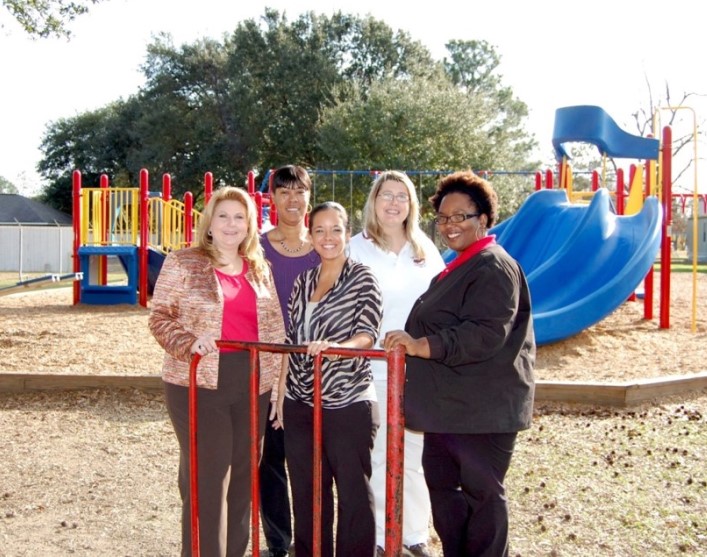 Five women administrators stand in front of a playground with one holding a red gate.