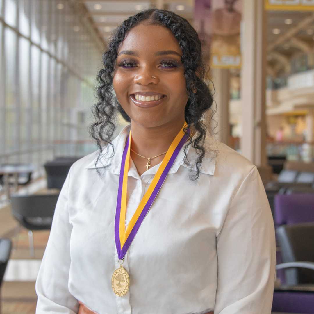Student in white blouse smiles with purple and gold ribbon and medal around neck.