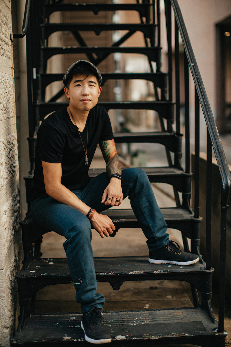Jo, a queer, trans nonbinary disabled Taiwanese American person, in a Black t-shirt and blue pants, seated on a metal staircase.