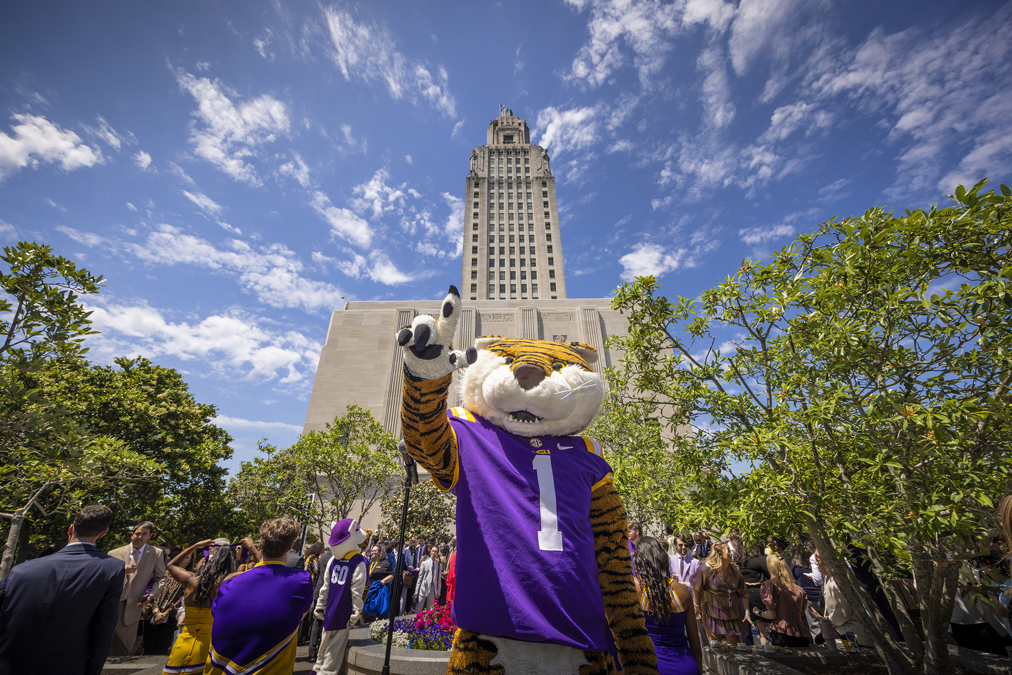 Mike the Tiger mascot, dressed in a purple #1 jersey, waves to the camera in front of the Louisiana State Capitol