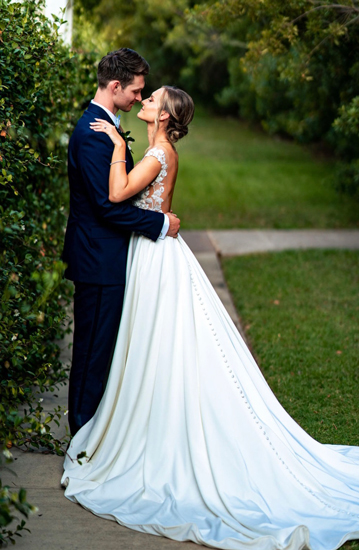 Kaleb Huckeba and Reka Perry embrace on their wedding day; the photo is taken in a lush green location and the couple is in wedding attire.