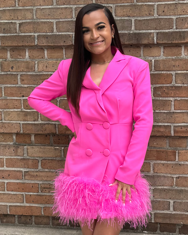 Cameron Beaugh wears a pink coat dress as she stands in front of a brick wall and smiles. 
