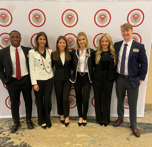 A group of six accounting majors in suits standing in front of a Beta Alpha Psi branded backdrop. 