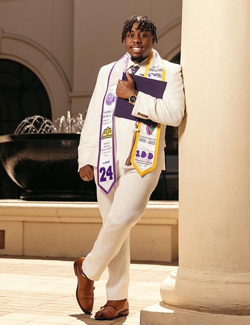 Chaderick Coleman wears a white suit and stoll from his undergraduate degree. The photo was taken on a sunny day by the foundation at the Lod Cook Alumni Center. 