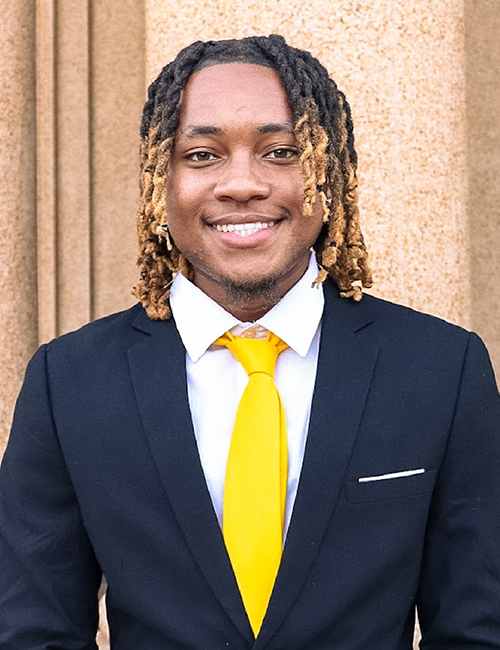Khalil Abdullah headshot wearing a dark suit and yellow tie. The photo is taken outdoors. Khalil has a warm smile. 
