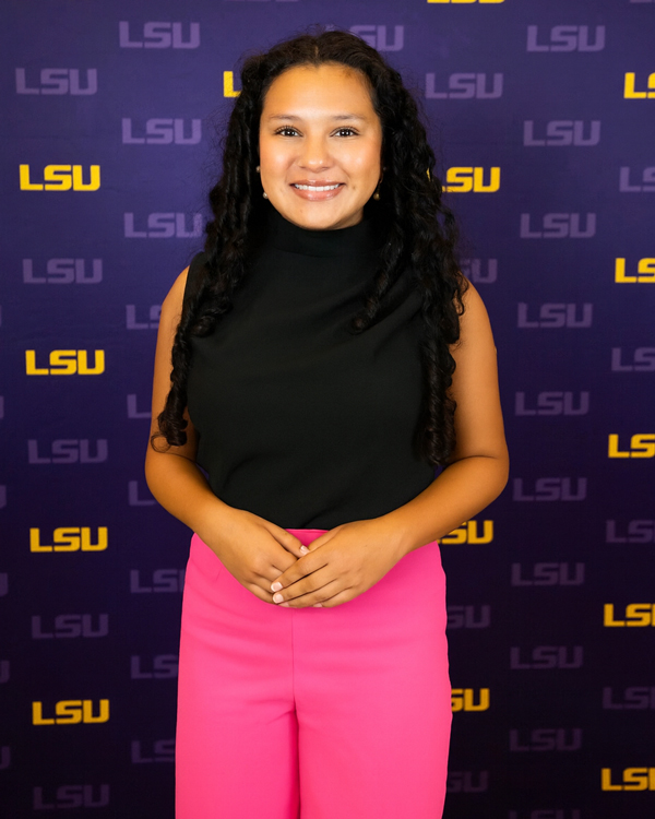 Maia Leonard stands in front of an LSU backdrop wearing a black blouse and pink pants. She smiles warmly.