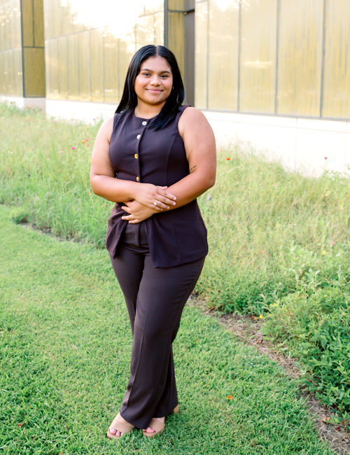 Marylet Morfin wears professional attire and smiles warmly in this photo taken at the Business Education Complex. In the background, the golden classroom wings can be seen with a few zinnia blooms popping out from the thick ornamental grass. 
