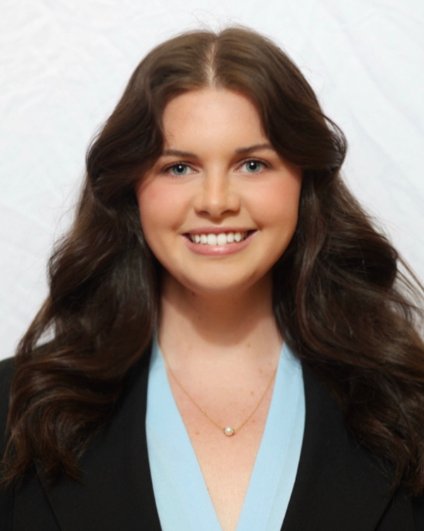 Claire Nickels wears a dark blazer and light blue blouse in this studio headshot. She smiles brightly.