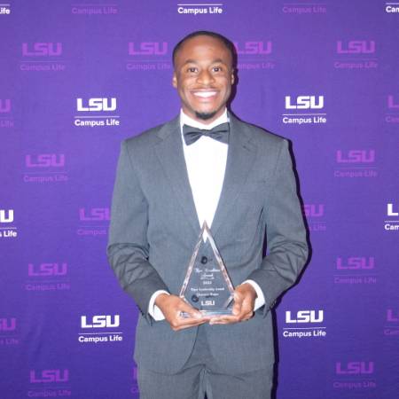 Clarence Magee II wears a tux and holds an award as he stands in front of a step and repeat with LSU branding.