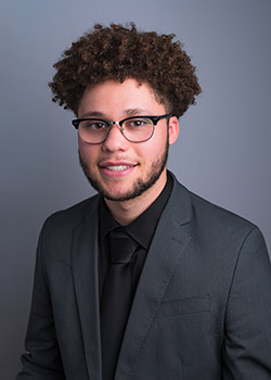 Jaydin Brewer's studio headshot, he wears a dark suit and glasses and smiles