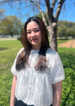 Thu Nguyen's headshot taken outside the Business Education Complex on a sunny day. She wears a fluid white blouse with dark polka dots.