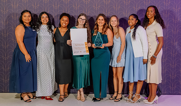 A group of women in dressy attire stand in front of a branded purple background and hold awards.