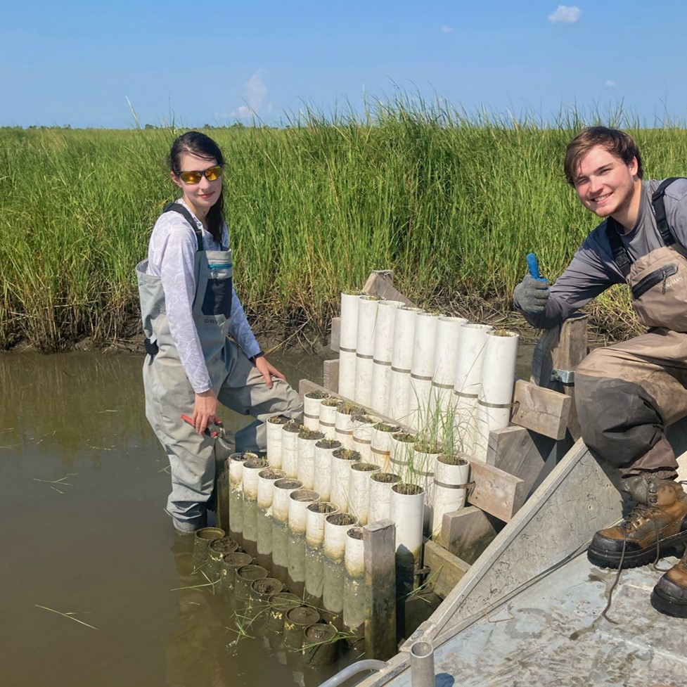 two people in waders pose with soil and vegetation samples in a marsh