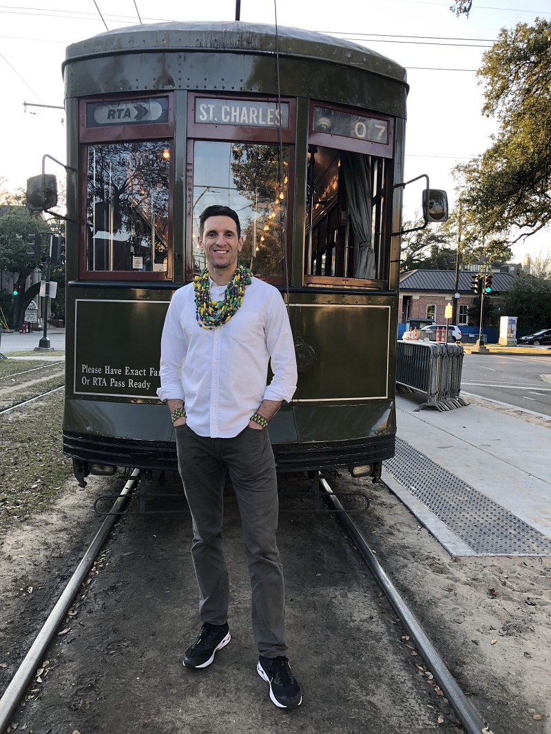 a man wearing Mardi Gras beads stands in front of a streetcar