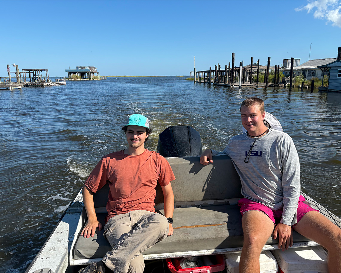 two men sit in the back of a boat on the water, with broken down cabins on stilts in the background