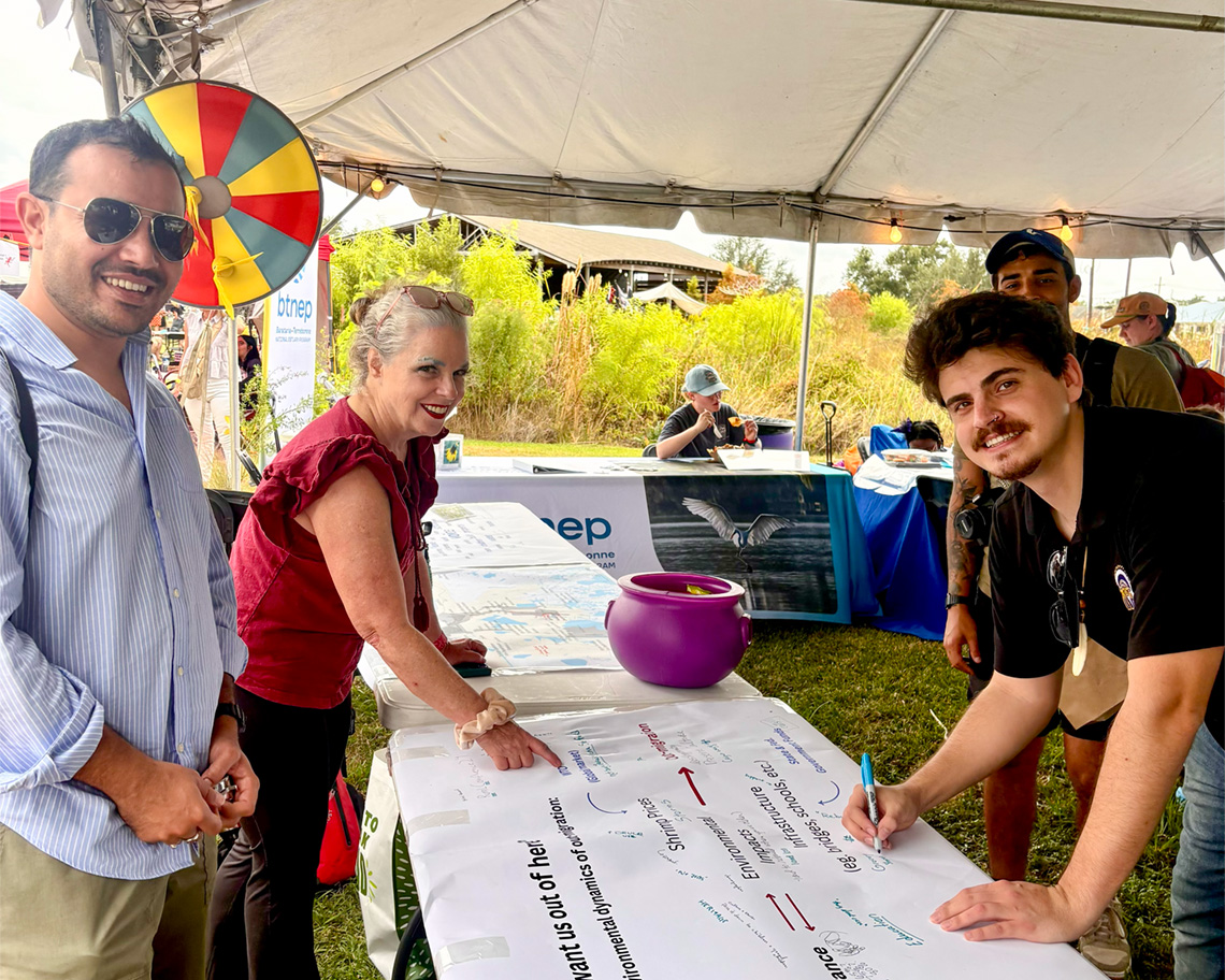 Three people stand around a table with a paper on it. One of them holds a pen.