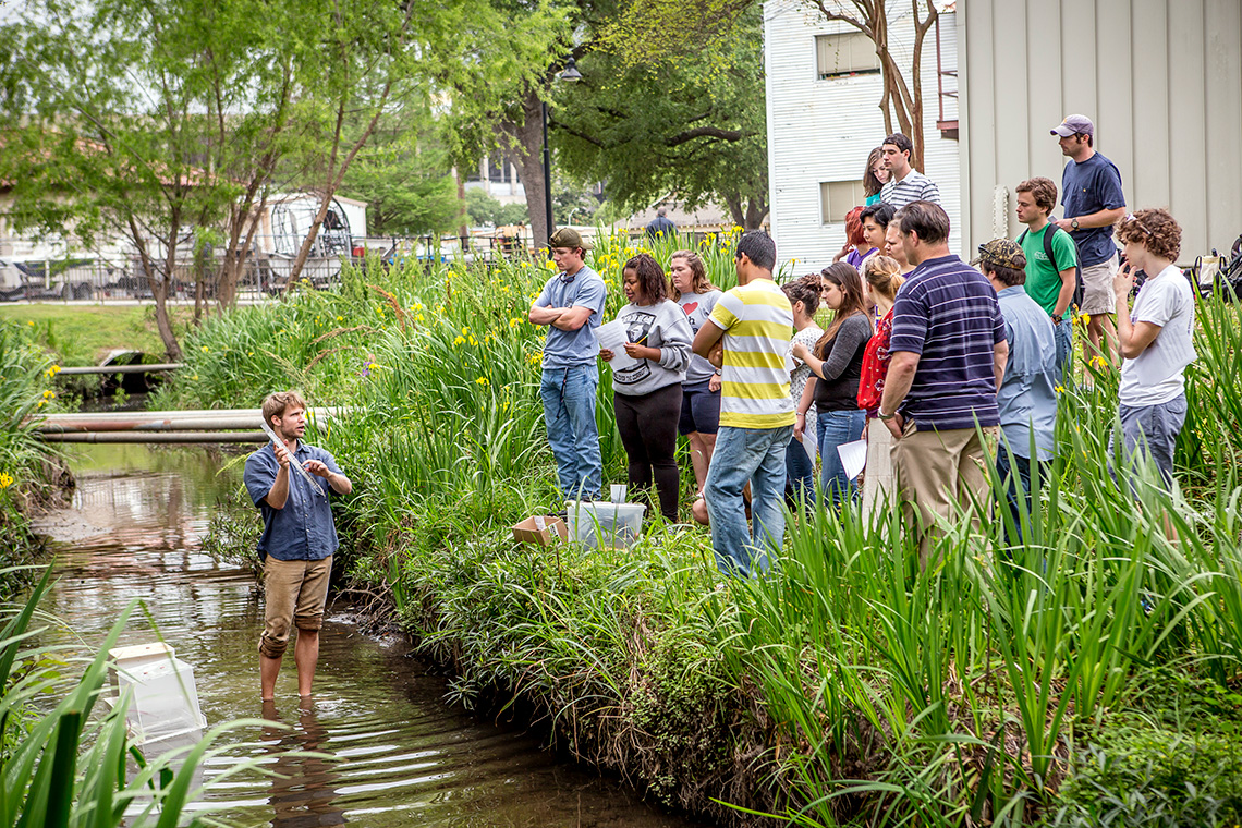 a group of students stand on the bank while a man in a river holds a test tube and talks