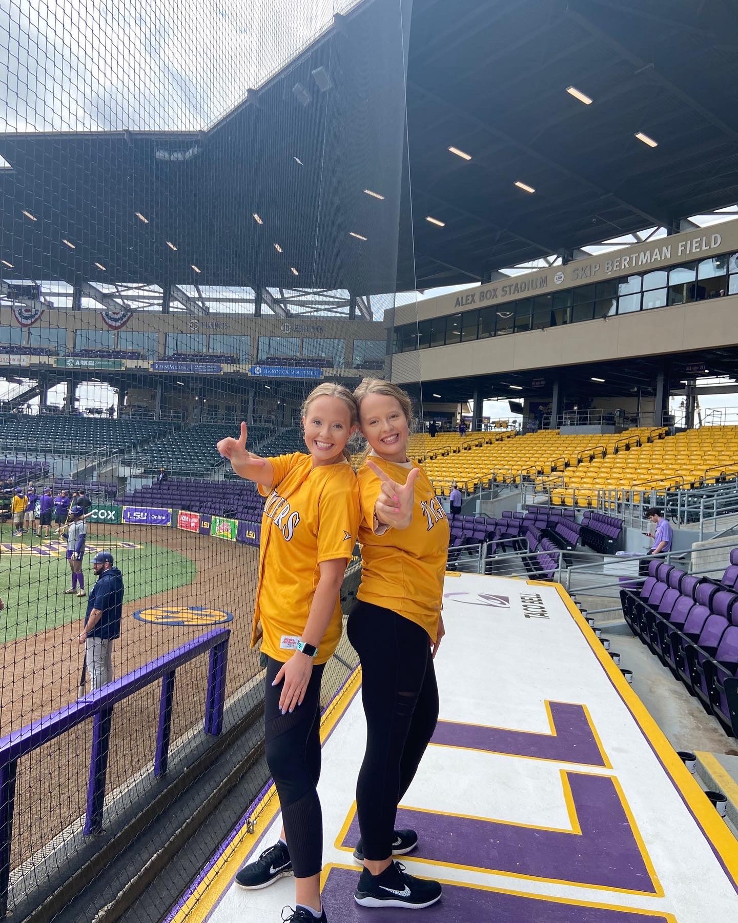 Two women stand back to back smiling. They both wear gold LSU jerseys