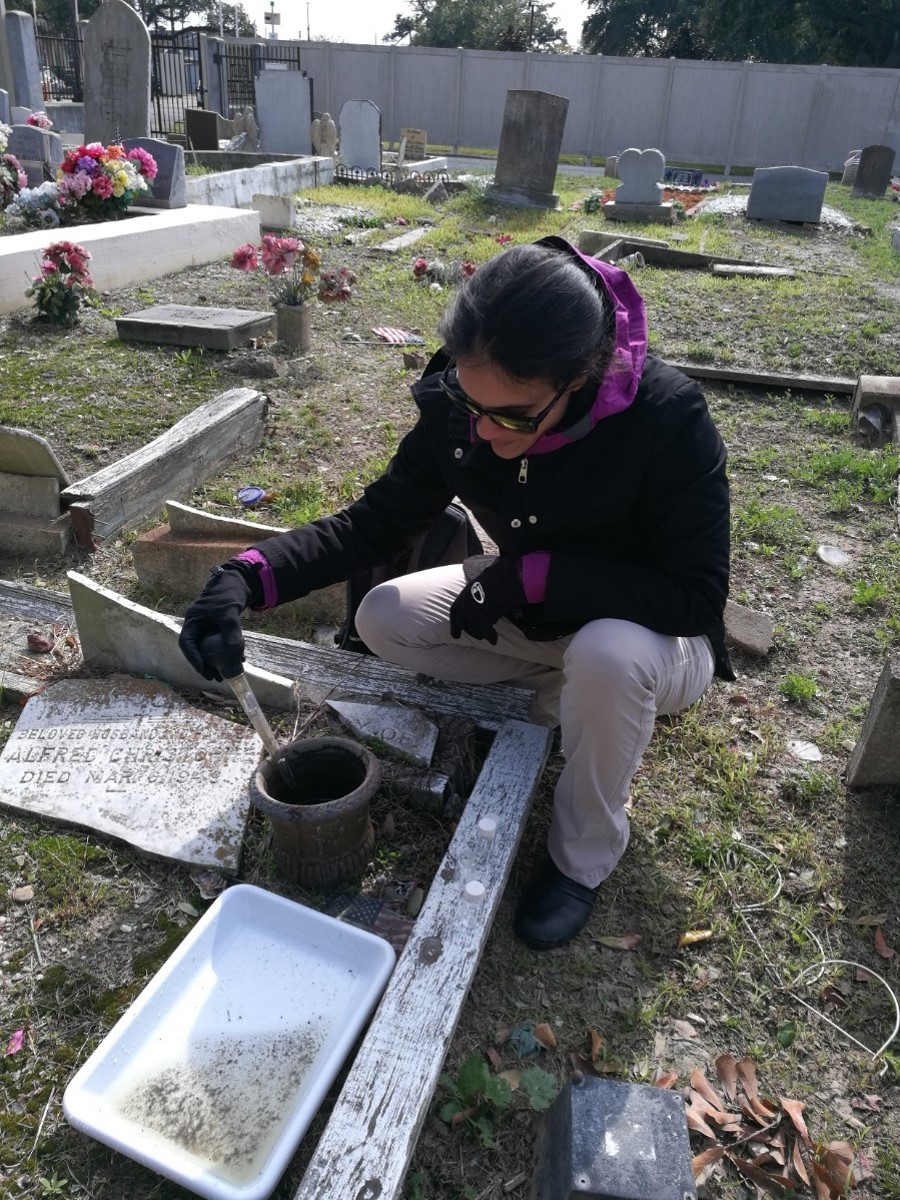 A woman kneels at a vase in a cemetary, using a large pipette to take a sample