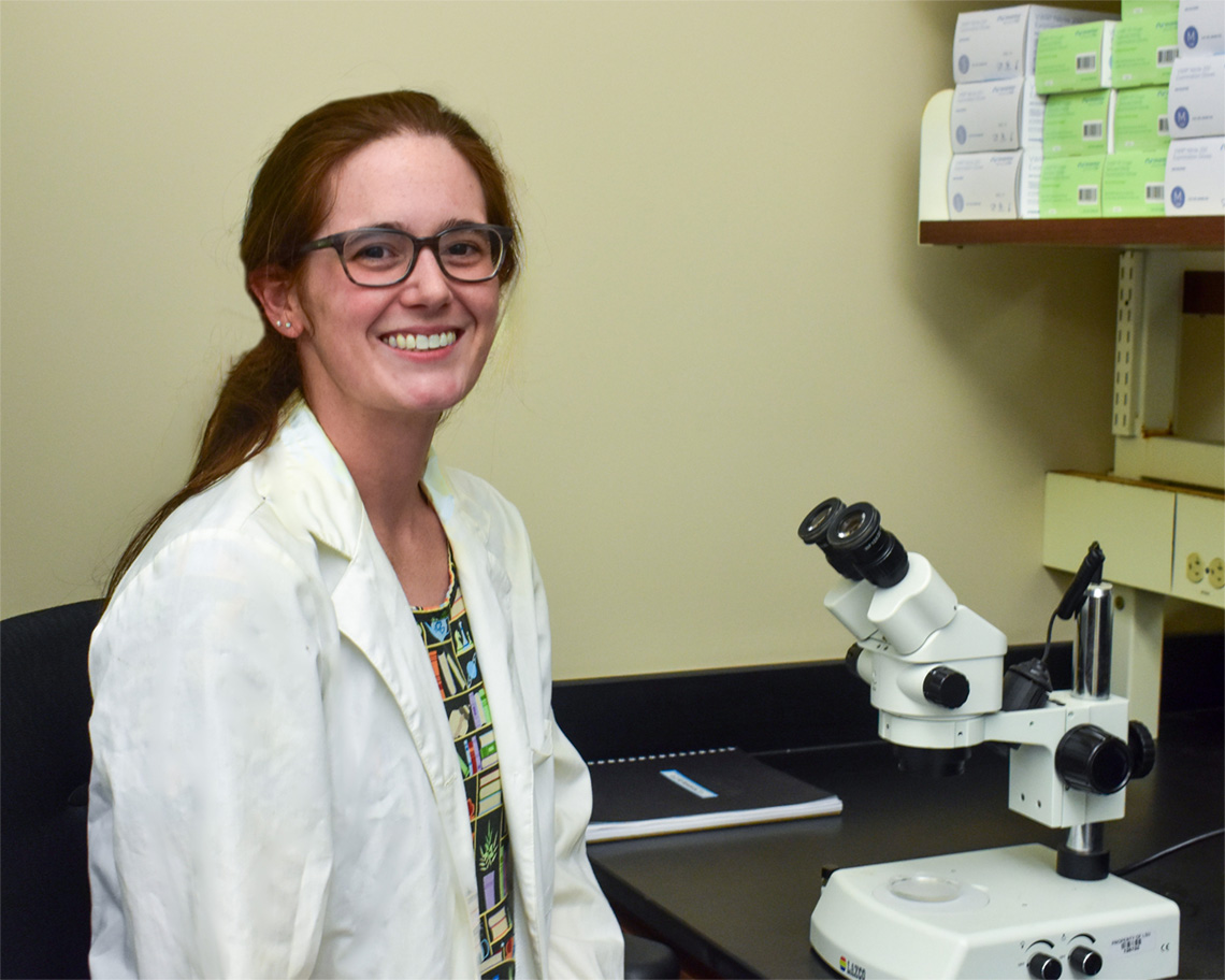 A woman in a lab coat sits next to a microscope