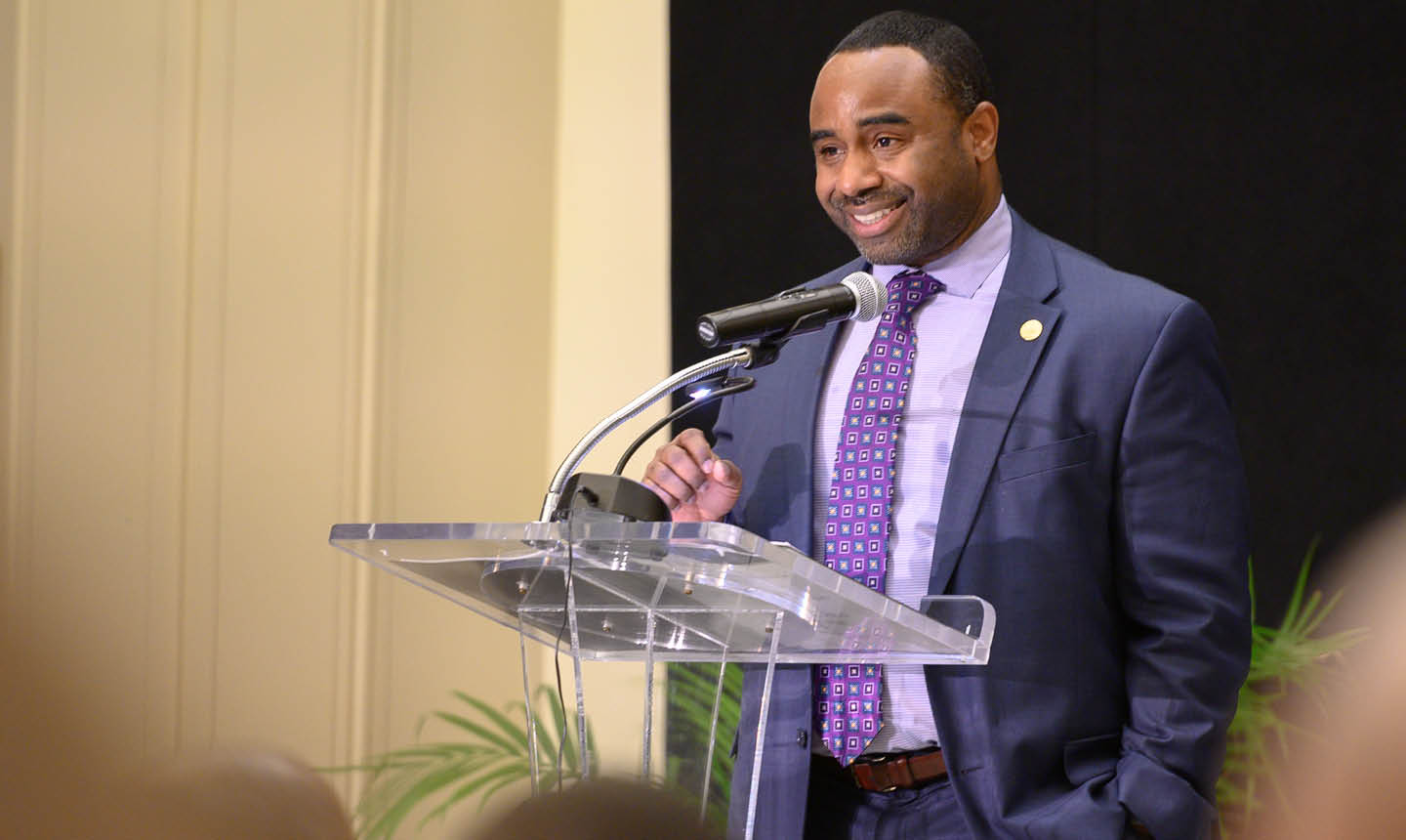 A person in a suit and patterned tie is speaking and smiling at a clear podium with a microphone in a formal indoor setting with an audience in the foreground. Description generated by AI