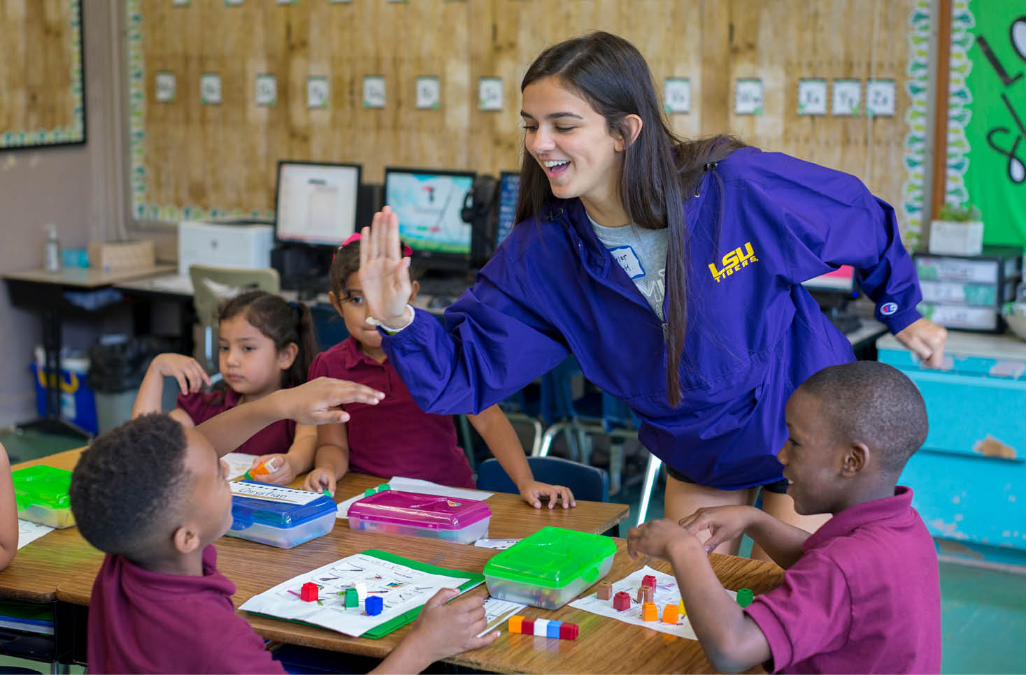 An adult wearing a purple LSU jacket is smiling and giving a high five to children seated at a classroom table engaged in an educational activity with colorful blocks. Description generated by AI