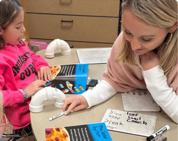 Three people sit at a table with books, a whiteboard with words written on it, and markers, engaged in a learning or teaching activity. Description generated by AI