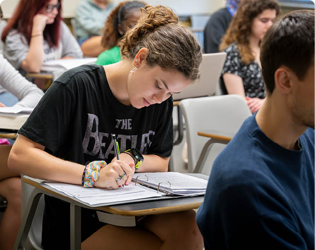 Three people sit at a table with books, a whiteboard with words written on it, and markers, engaged in a learning or teaching activity. Description generated by AI