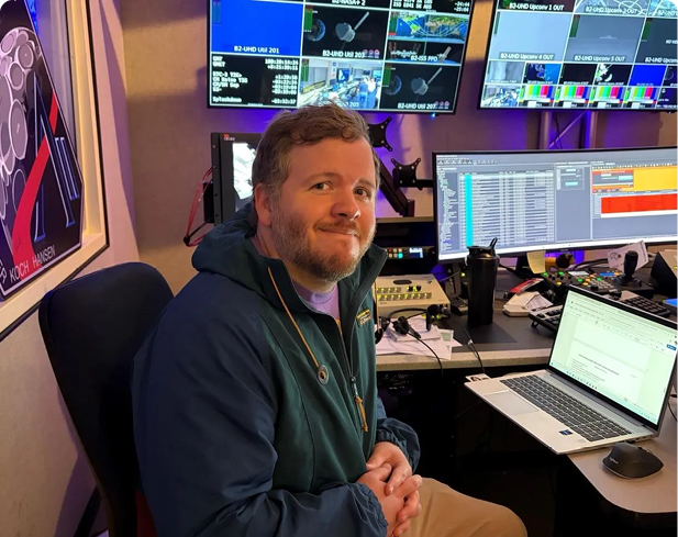 A person with short curly hair and a beard is smiling while sitting in front of multiple computer monitors displaying various data and colorful screens in a dimly lit room. Description generated by AI
