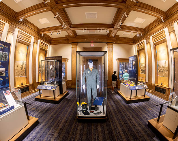 A museum room with wooden paneling and display cases featuring historical clothing and informational panels, with two people observing the exhibits. Description generated by AI