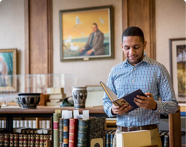 A person wearing a checkered shirt is reading a book in a room decorated with framed paintings and pottery. Description generated by AI