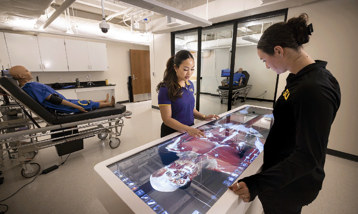 Two adults interact with a large touchscreen table displaying a detailed anatomical image while a person lies on a medical bed in a clinical setting. Description generated by AI