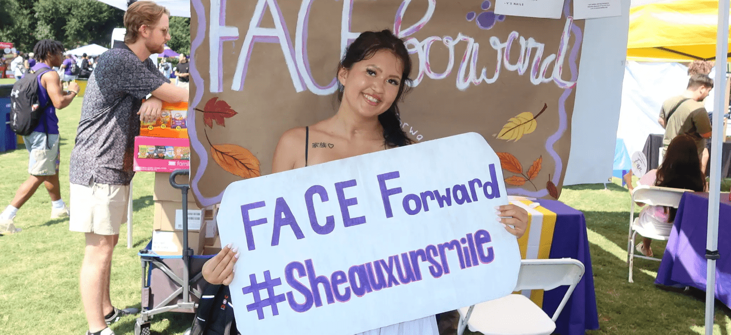 A person with long hair is smiling and holding a sign that says “FACE Forward #Sheauxursmile" at an outdoor event with other people and booths in the background. Description generated by AI