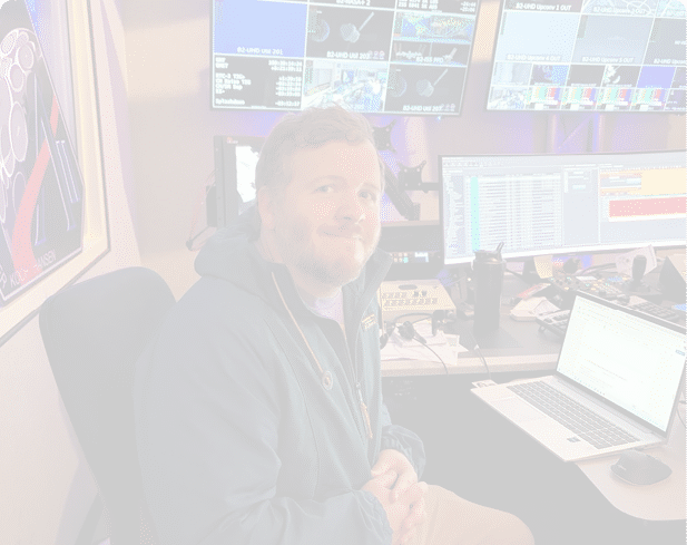 A person with short curly hair and a beard is smiling while sitting in front of multiple computer monitors displaying various data and colorful screens in a dimly lit room. Description generated by AI