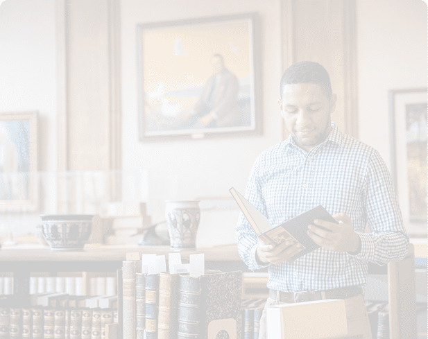 A person wearing a checkered shirt is reading a book in a room decorated with framed paintings and pottery. Description generated by AI