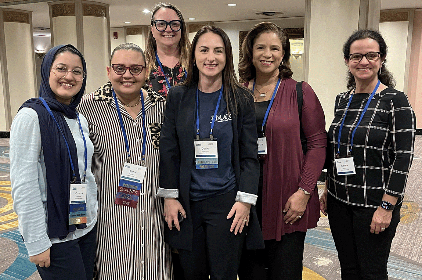 Six adults wearing conference badges stand closely together indoors smiling and posing for a group photo. Description generated by AI