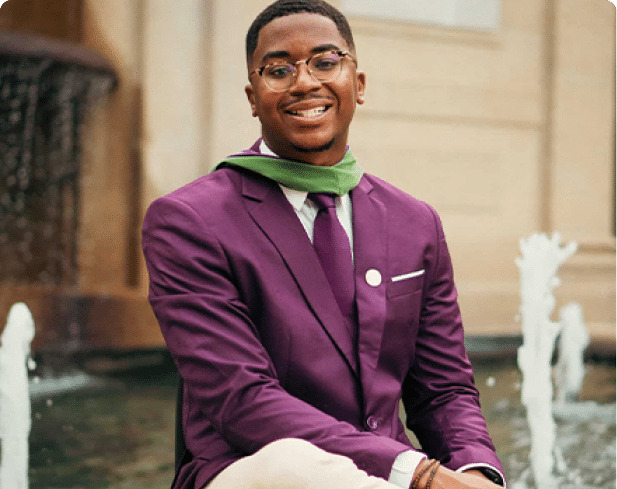 A person wearing a purple blazer, beige pants, and a tie is sitting cross-legged near a water fountain. Description generated by AI