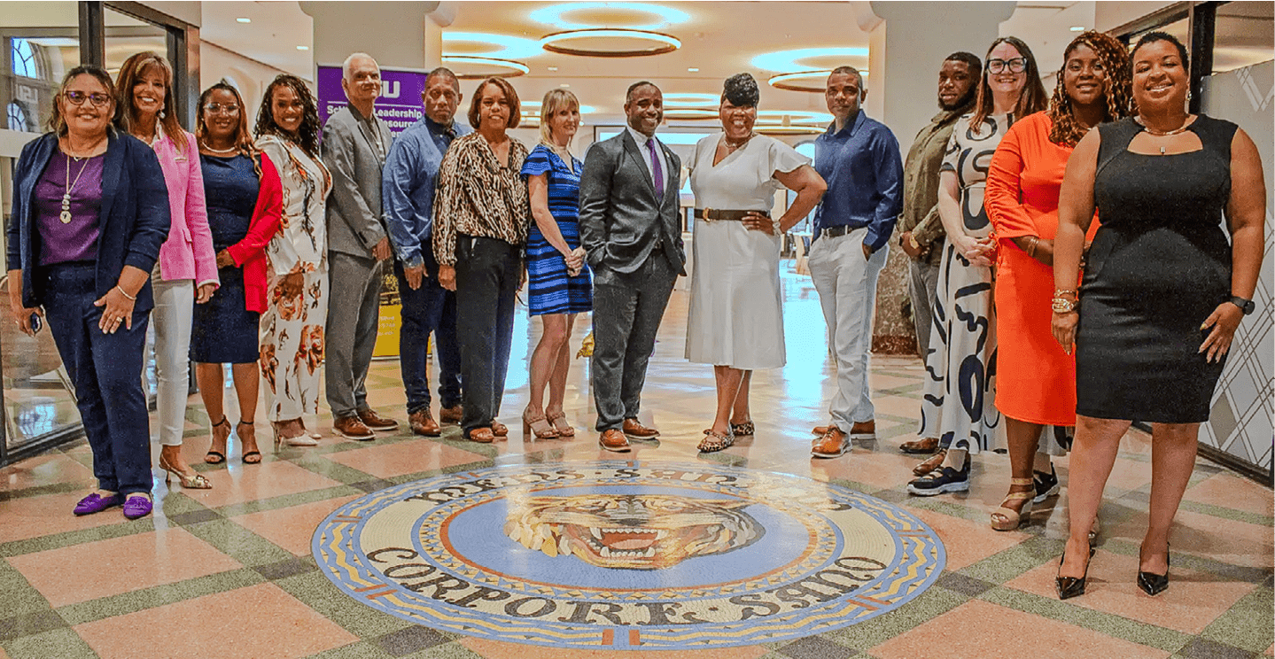 A diverse group of people dressed in business and formal attire stand together indoors around a circular emblem on the floor in a well-lit space. Description generated by AI