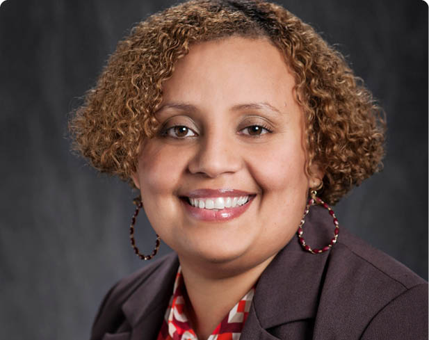 A person with curly hair is smiling and wearing a patterned red and white blouse under a dark blazer with large hoop earrings against a dark background. Description generated by AI