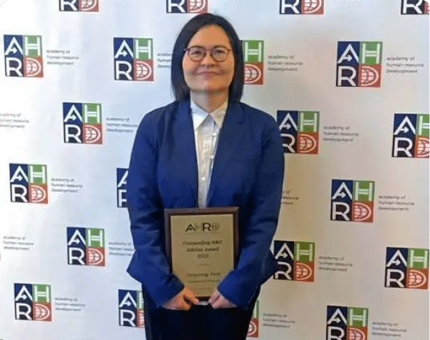A person with shoulder-length hair and glasses is smiling while wearing a blue blazer and white collared shirt in front of a backdrop with logos and text. Description generated by AI
