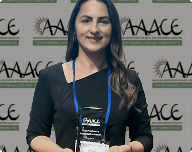 A person with long wavy hair wearing a black top and a blue lanyard is smiling in front of a backdrop with repeated logos and text. Description generated by AI