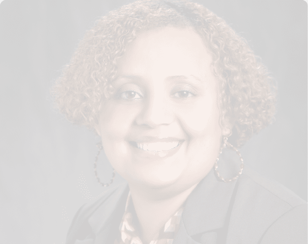 A person with curly hair is smiling and wearing a patterned red and white blouse under a dark blazer with large hoop earrings against a dark background. Description generated by AI