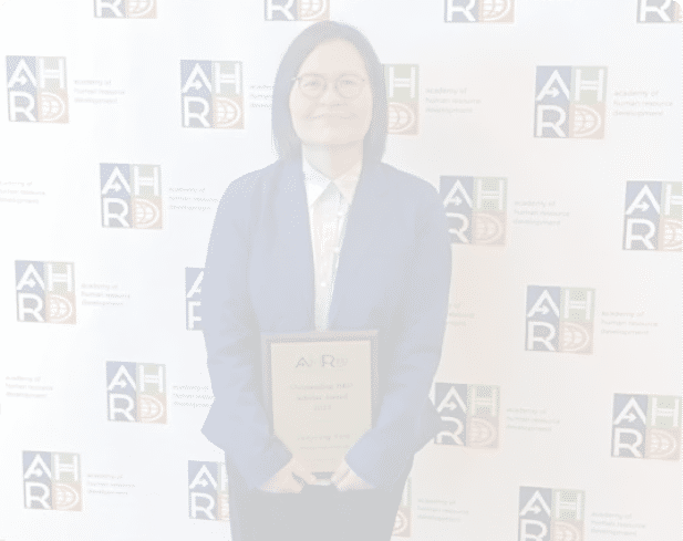 A person with shoulder-length hair and glasses is smiling while wearing a blue blazer and white collared shirt in front of a backdrop with logos and text. Description generated by AI