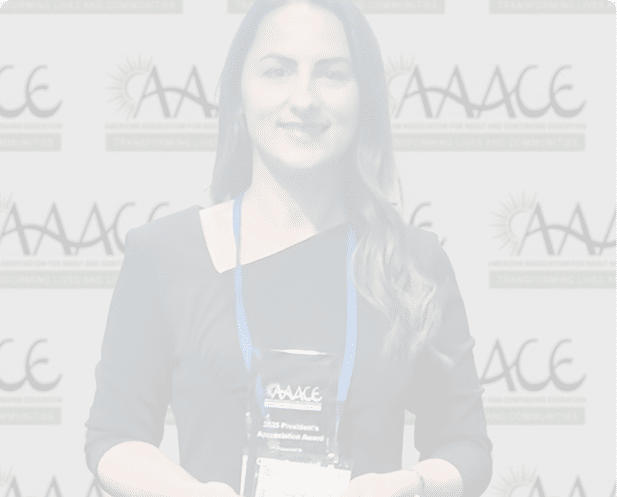 A person with long wavy hair wearing a black top and a blue lanyard is smiling in front of a backdrop with repeated logos and text. Description generated by AI