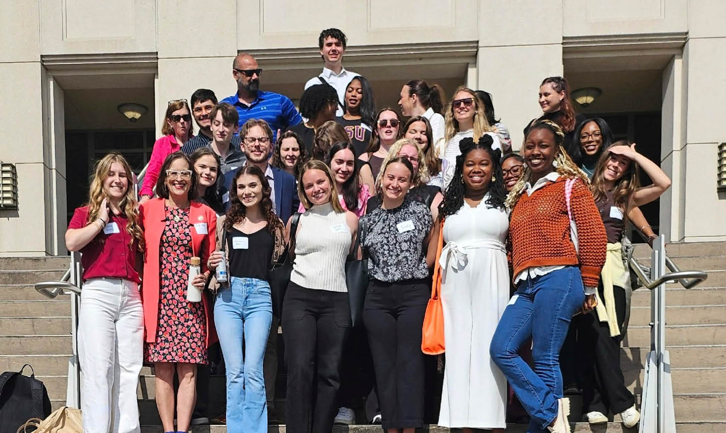 A group of adults smiling and posing together on outdoor steps in casual and semi-formal clothing on a sunny day. Description generated by AI