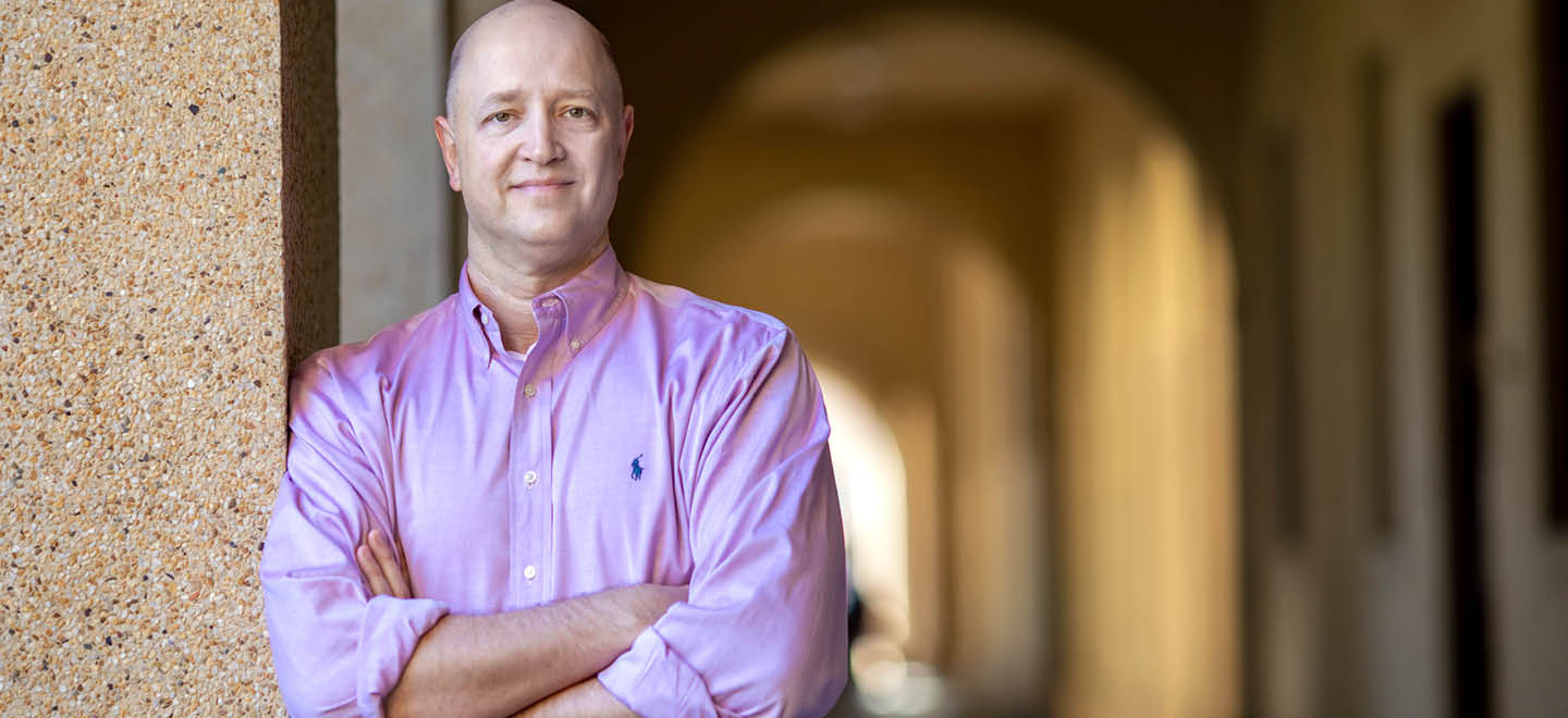 A person wearing a light purple button-up shirt with rolled-up sleeves is standing with arms crossed and leaning against a textured wall in a corridor with arched doorways. Description generated by AI